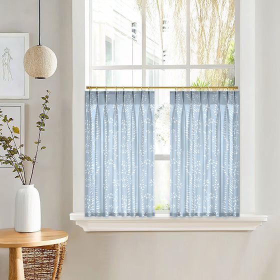 Light blue curtarra custom curtains with a subtle pattern hanging in a window, with a white vase and plant on a wooden table to the left.