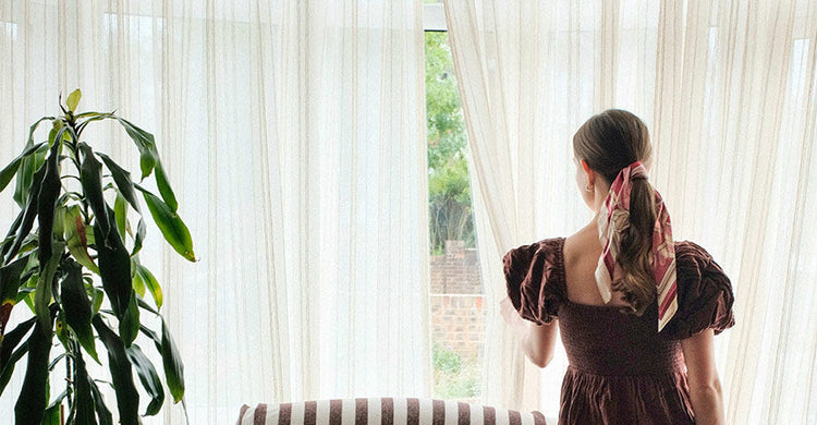 Sheer cream curtains filtering light in a cozy home, a woman looking outside. Window treatments for cozy living.