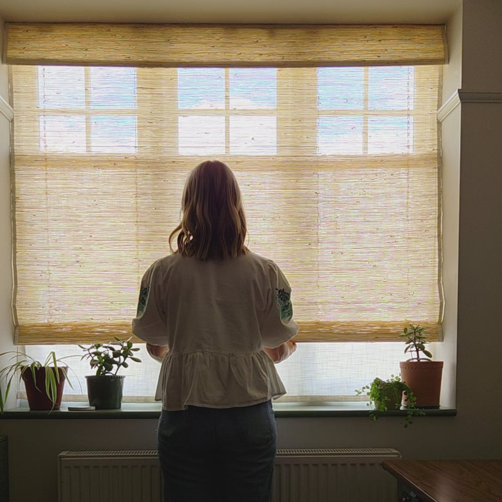 Pale Mimosa,Woman standing by a window with a natural woven wood shade, showcasing its light-filtering qualities and organic aesthetic.